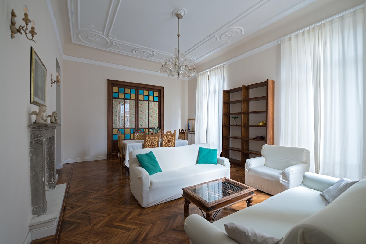 A bright living room features high ceilings with ornate details and large windows dressed in sheer white curtains. Two white sofas are arranged with teal cushions, positioned around a wooden coffee table. A wooden bookshelf stands against the wall, and a dining area is visible in the background.