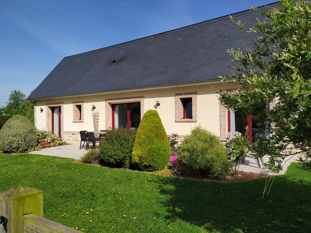 The exterior of the renovated farmhouse features a light-colored façade with red window frames. A spacious patio area includes outdoor seating. Lush greenery and ornamental shrubs surround the property, enhancing the peaceful ambiance. The roof is a dark shade, contrasting with the afternoon sky.