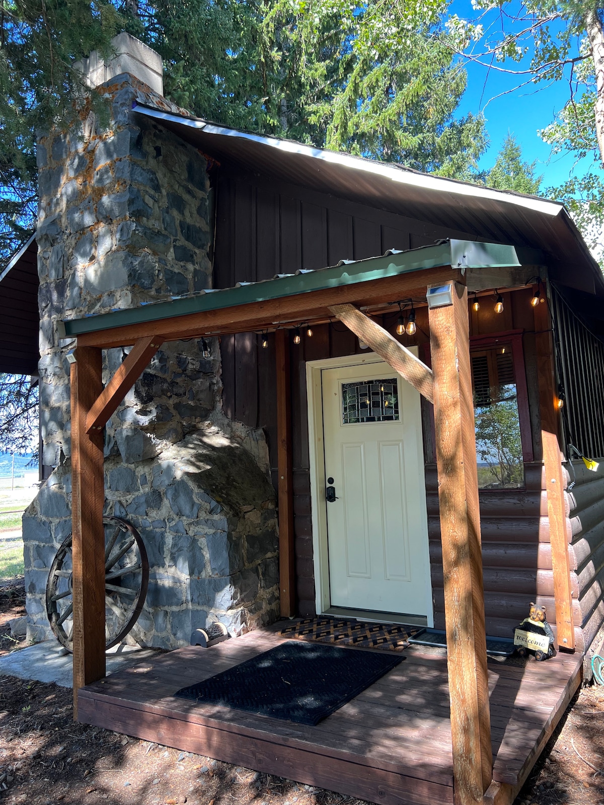 The exterior of the cabin features a large stone fireplace and a welcoming entrance with a white door. A covered porch area is adorned with string lights, adding a cozy touch. Lush green trees surround the cabin, creating a serene setting.
