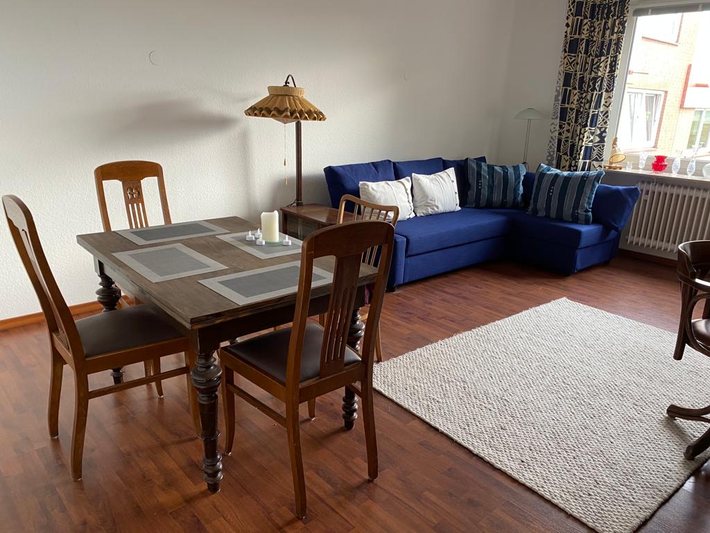 A dining area features a wooden table surrounded by four chairs, with a candle and placemats arranged on top. In the living space, a blue sectional sofa offers comfortable seating, accented by decorative pillows. A large window lets in natural light, illuminating the space.