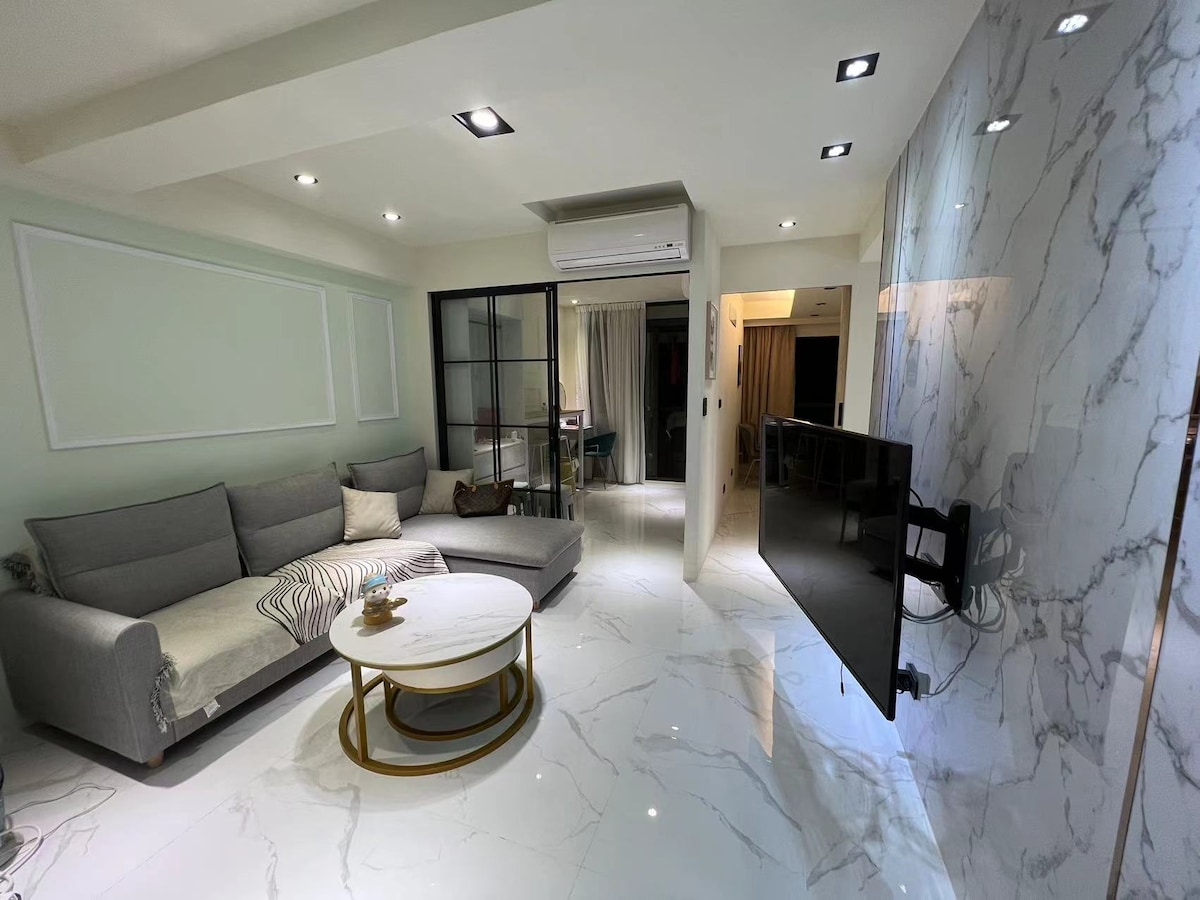 A spacious living room is presented with a comfortable sectional couch and a round coffee table. A large flat-screen TV is mounted on a sleek, marbled wall. Natural light enhances the open layout, leading to a glass door that connects to the rest of the apartment.