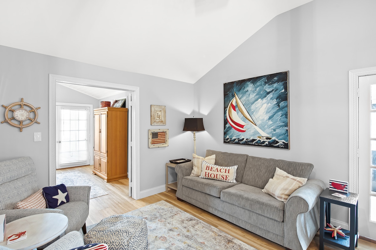 A light-filled living area features a gray sectional sofa adorned with beach-themed pillows. A large painting of a sailboat decorates the wall, while a lamp stands beside the couch. Wood accents and a textured rug add warmth to the space, enhancing its coastal charm.