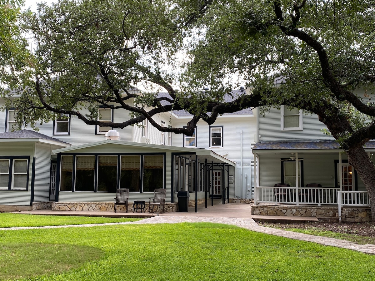 The image captures the exterior of a multi-story inn surrounded by lush green grass and trees. A spacious patio area features outdoor seating, while large windows offer glimpses of the inviting interior. The serene setting is complemented by a gentle canopy of overhanging branches.