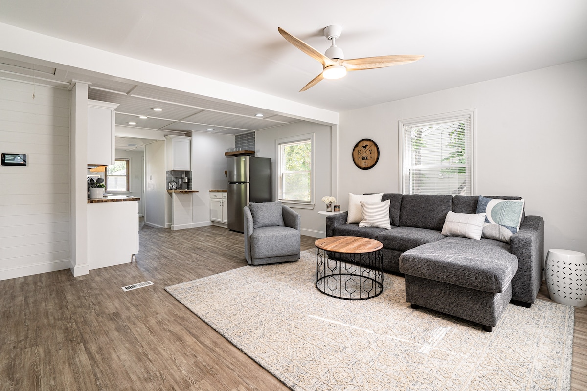 A modern living area is showcased, featuring a comfortable sectional sofa and a round coffee table. Natural light filters through large windows, illuminating the space with soft brightness. A ceiling fan enhances air circulation, while a patterned area rug adds texture to the floor.