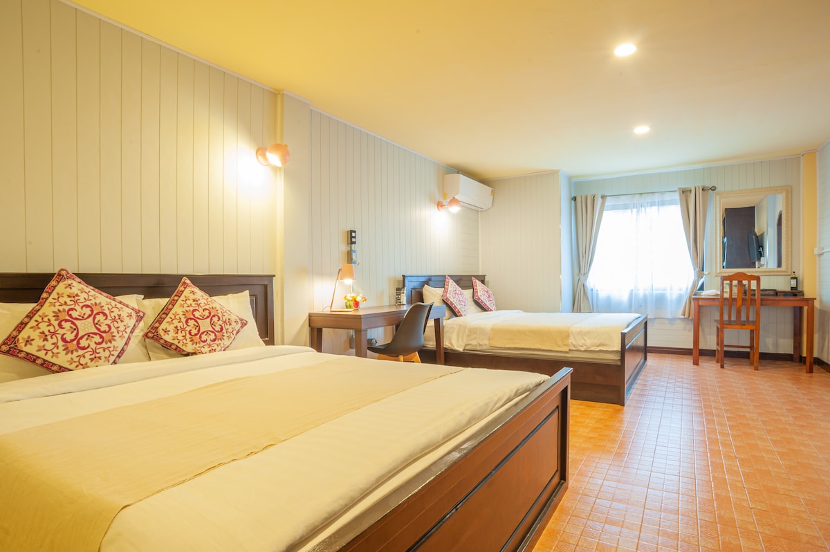 A spacious guest room features two beds with decorative pillows and an inviting atmosphere. A work desk is positioned near a window, allowing natural light to brighten the space. Soft lighting enhances the warm tones of the room, complementing the cozy furnishings.