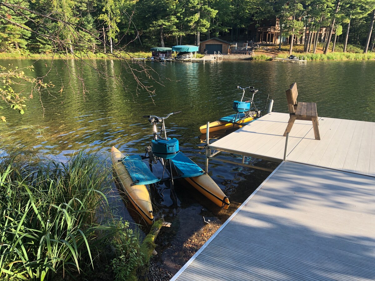 Two colorful paddle bikes are positioned on a private dock, with scenic views of the tranquil lake and surrounding trees. The calm water reflects the greenery, while a wooden chair is placed nearby for relaxing by the water's edge.