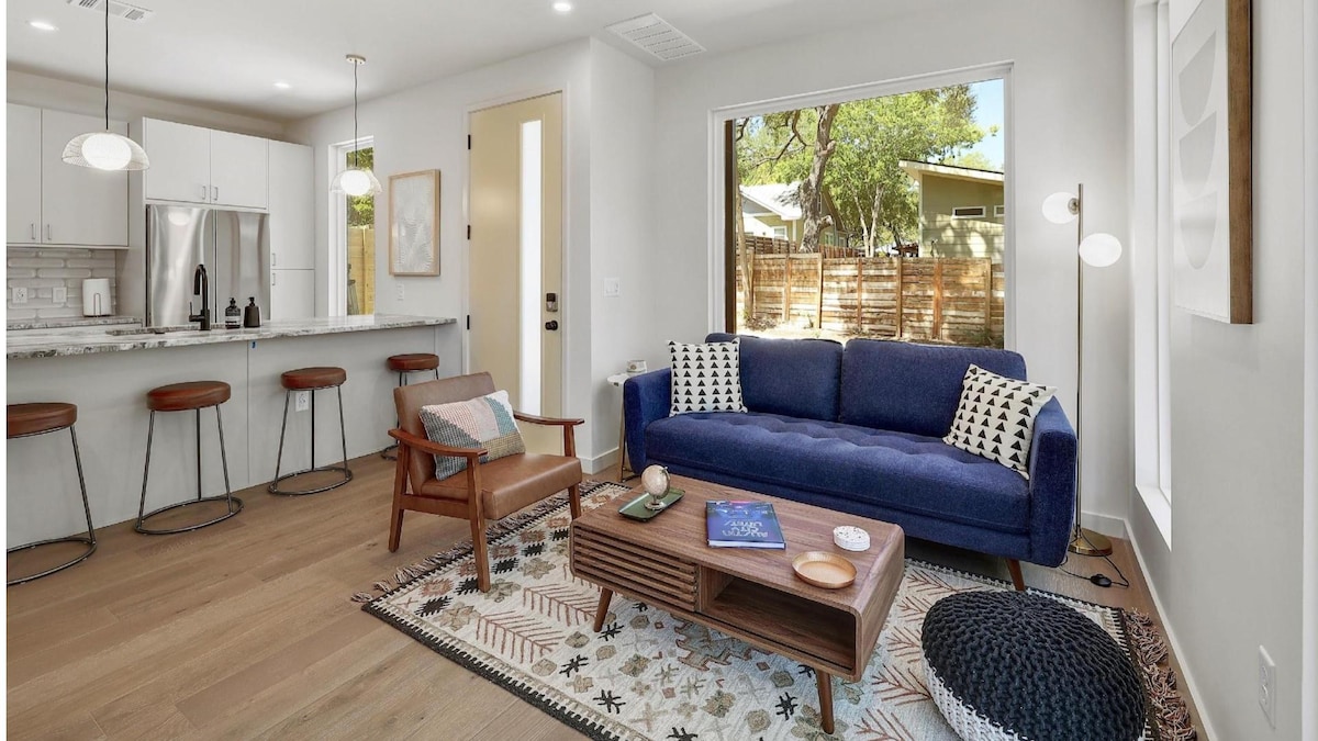 A modern living area features a deep blue sofa with decorative cushions positioned against a window that overlooks a private fenced yard. A wooden coffee table is centered on a patterned area rug, complemented by a natural wood armchair and bar stools at a nearby kitchen counter.