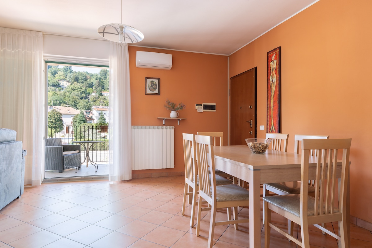 A dining area is presented with a light wooden table surrounded by six chairs. An inviting orange wall provides a warm backdrop, while a large window opens to a view of the outdoors. A decorative piece adds interest to the space.