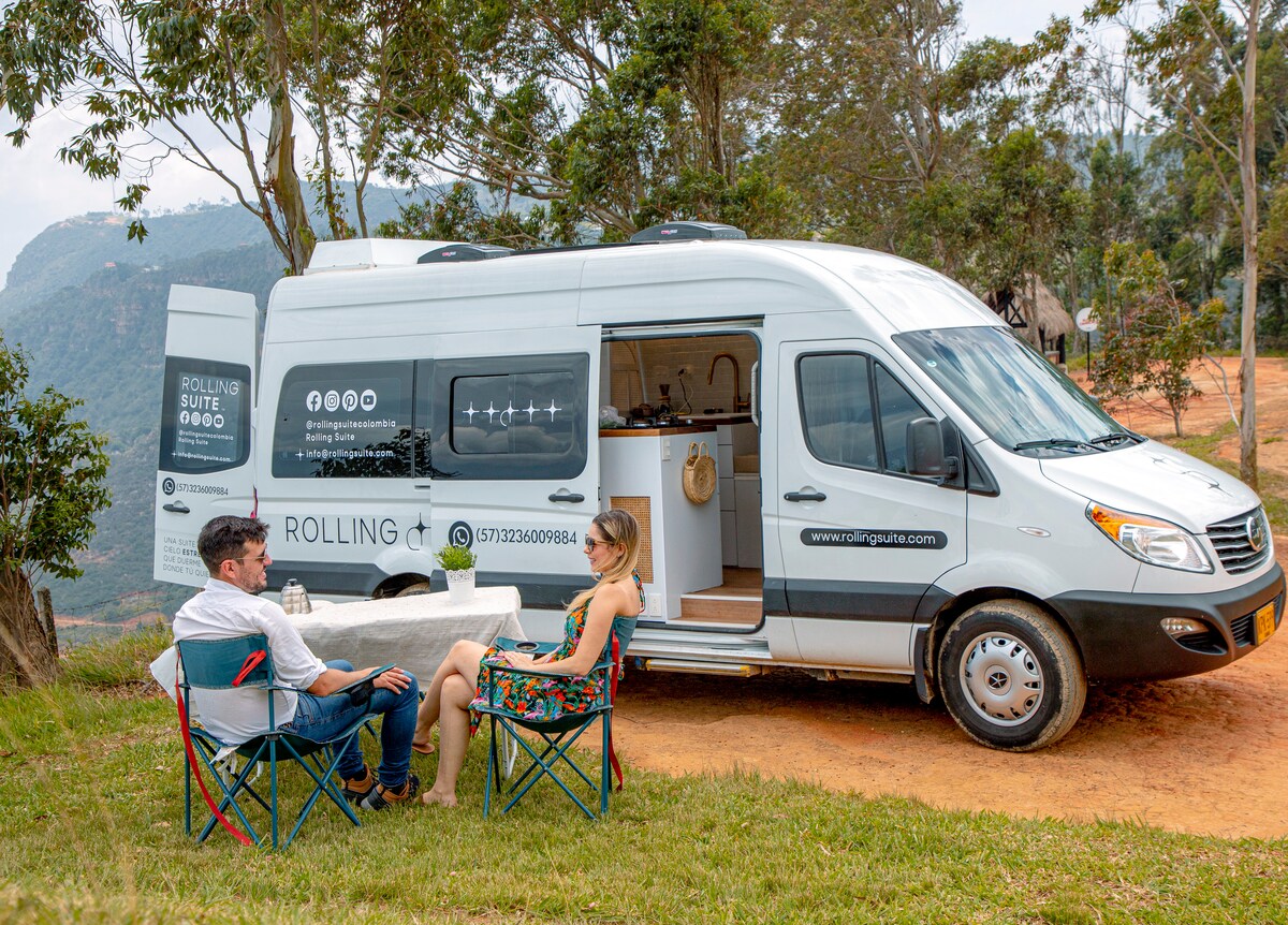 A cozy outdoor dining setup is displayed next to a stylish camper van. Two folding chairs are positioned around a small table, offering a serene view of rolling hills in the background. The camper van, featuring a sleek design and modern amenities, is parked on a grassy area.