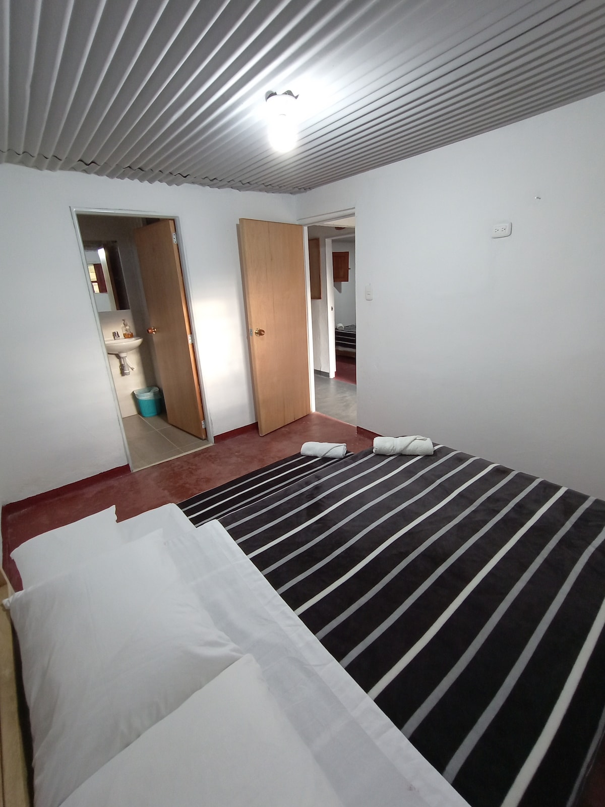 A sleeping area is defined by a striped black and white blanket over a spacious bed, with two rolled towels placed neatly on top. A doorway to the bathroom is visible, along with a glimpse of light coming from the room beyond.