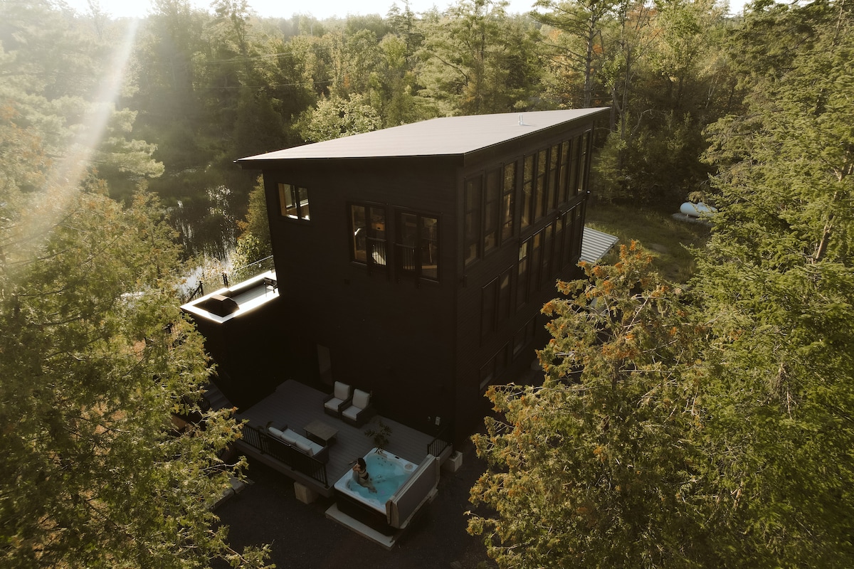 An aerial view of the modern lake house reveals its sleek black exterior surrounded by lush greenery. A spacious patio is visible, featuring a hot tub and comfortable seating, set against the backdrop of trees and the serene lake beyond.