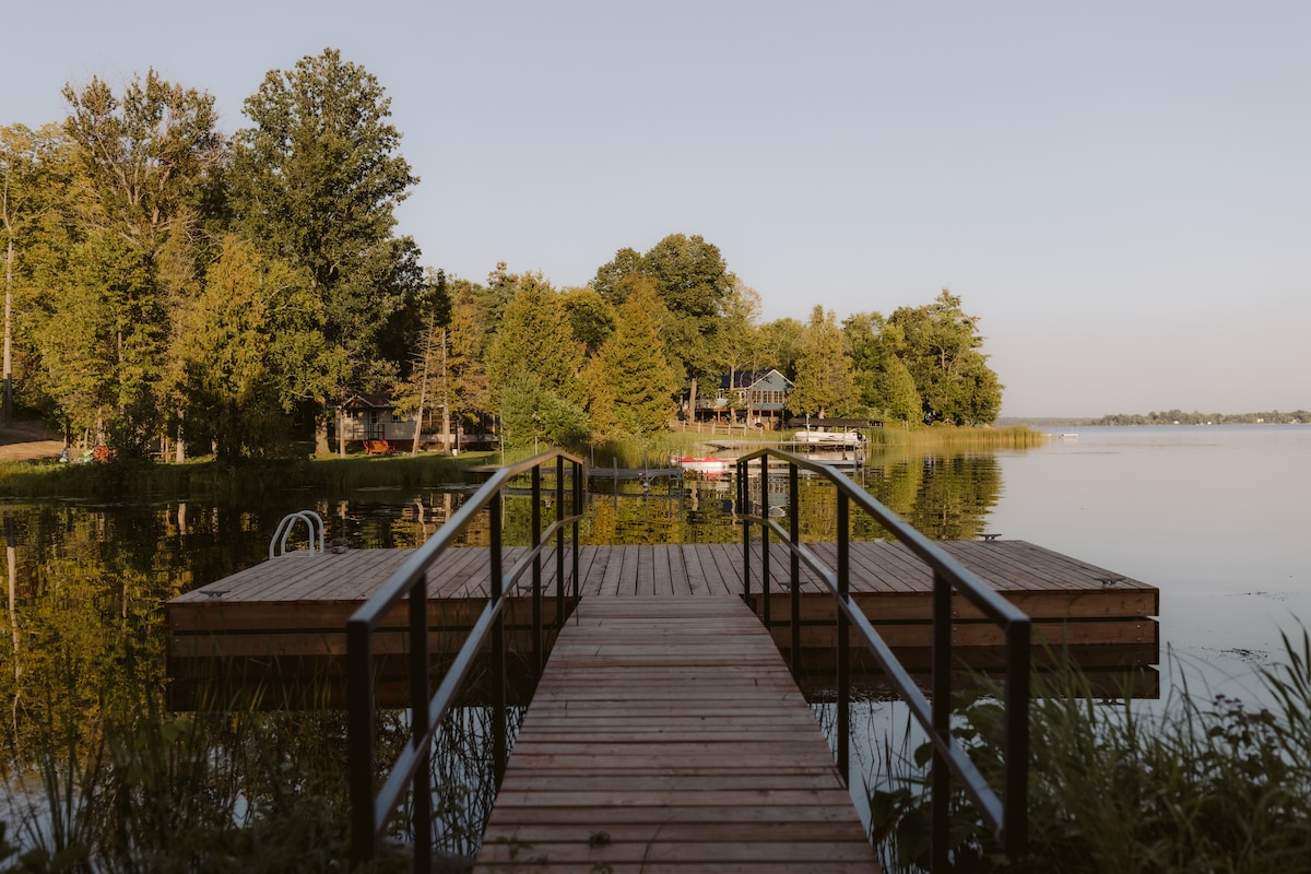 A wooden dock extends over calm waters, leading to a serene lakeside. Lush trees frame the shoreline, while neighboring structures are faintly visible across the water. The tranquil scene is enhanced by the soft light of early evening.