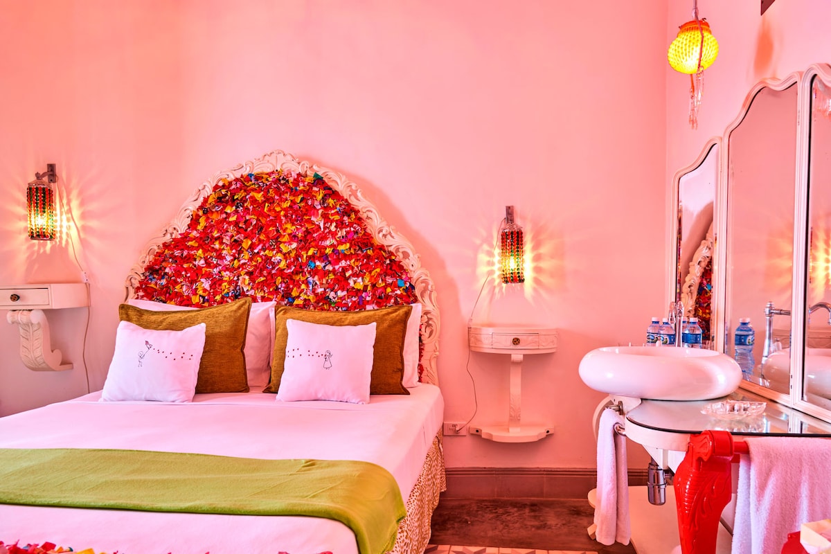 A vibrant bedroom features a headboard adorned with colorful flowers, complemented by two plush pillows. The white bedspread contrasts with the green accent blanket. A stylish double sink with a modern faucet is visible, along with decorative lighting fixtures that enhance the ambiance.
