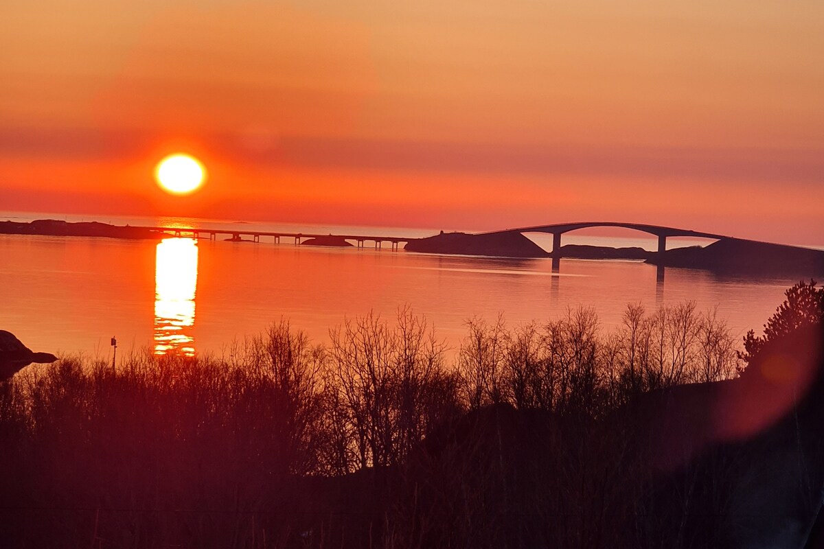 A serene sunset is reflected on calm waters, with a bridge silhouetted against the vibrant orange and pink sky. The surrounding landscape features soft silhouettes of trees, enhancing the tranquil atmosphere of the scene.