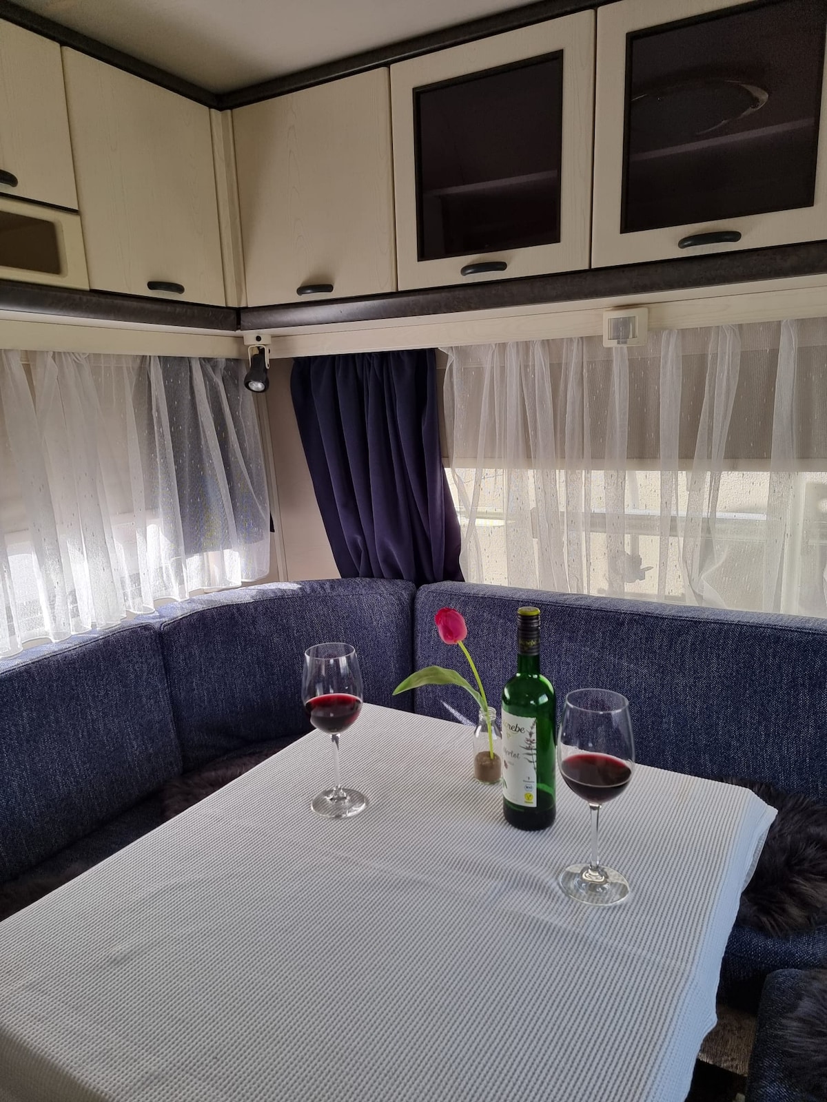 A dining area within the caravan features a white tablecloth covering a table, complemented by two glasses of red wine and a bottle. Natural light filters through sheer curtains, enhancing the cozy atmosphere created by the blue upholstery and cabinetry.