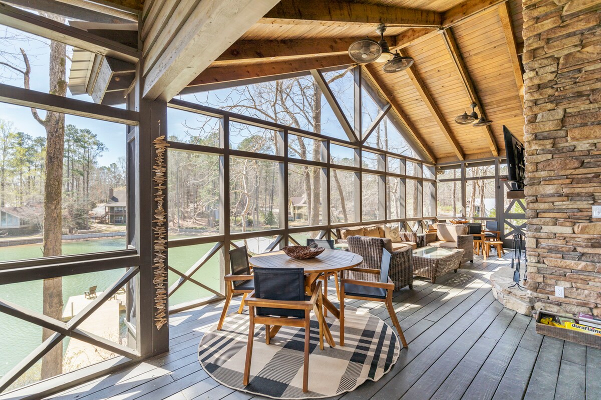 A screened sitting area offers views of the lake, featuring a round wooden table surrounded by chairs. Comfortable seating is arranged with cushions, and a stone fireplace is visible. Natural light filters through the windows, enhancing the serene atmosphere of the lakeside retreat.