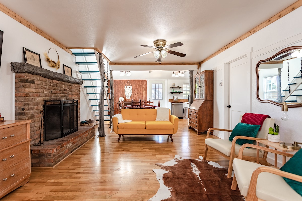 An open living area features a warm wood floor, highlighted by a brick fireplace flanked by modern furniture. A sunny yellow couch sits in the center, accompanied by two armchairs. The space is illuminated by ceiling fans and contains a staircase leading to the upper level.