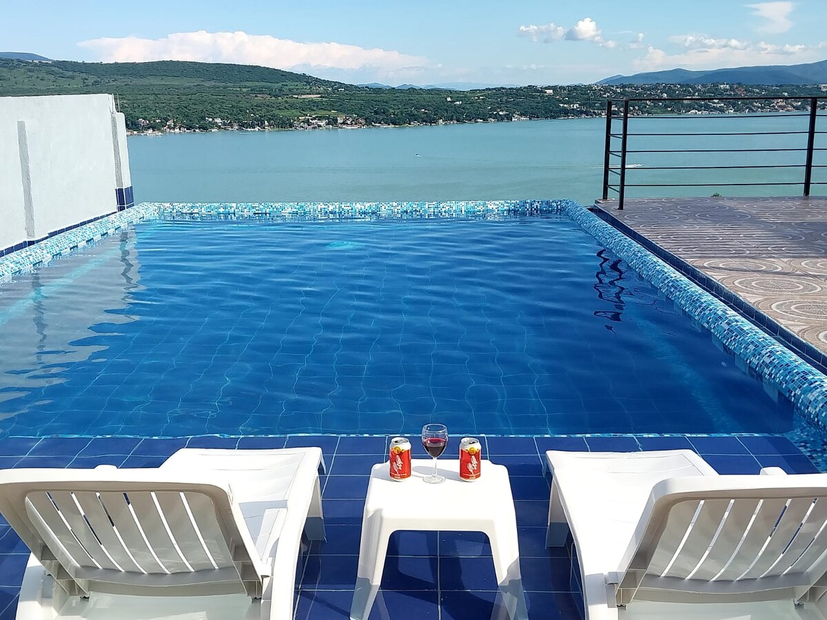 An infinity pool stretches across the foreground, reflecting the sky and offering views of the lake. Two lounge chairs face the water, with beverages placed on small tables nearby. The surrounding area features a tiled deck, contributing to a serene outdoor setting.