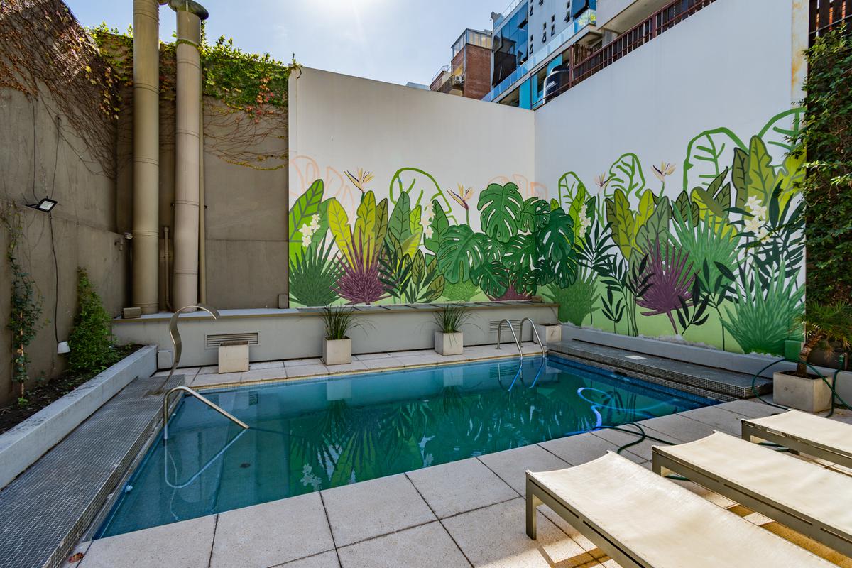 A serene pool area is framed by vibrant green murals depicting lush foliage, creating a refreshing environment. Sun loungers are positioned on the tiled deck, inviting relaxation. Natural light illuminates the water's surface, reflecting the surrounding greenery and the clear blue sky above.