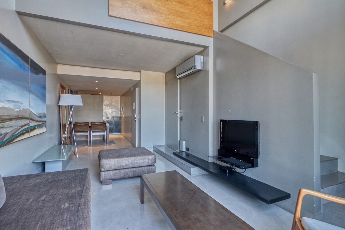 An open living area features a modern design with a neutral color palette. A comfortable sofa is positioned next to a sleek coffee table. A flat-screen television is mounted on the wall, and a dining area is visible in the background, enhancing the functional layout.
