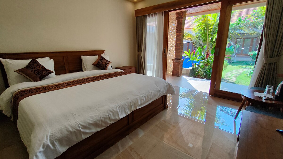 A spacious bedroom features a large, inviting bed with decorative pillows. Floor-to-ceiling glass doors lead to a serene outdoor garden area, allowing natural light to fill the room. A small wooden desk adds functionality, enhancing the overall relaxing ambiance.