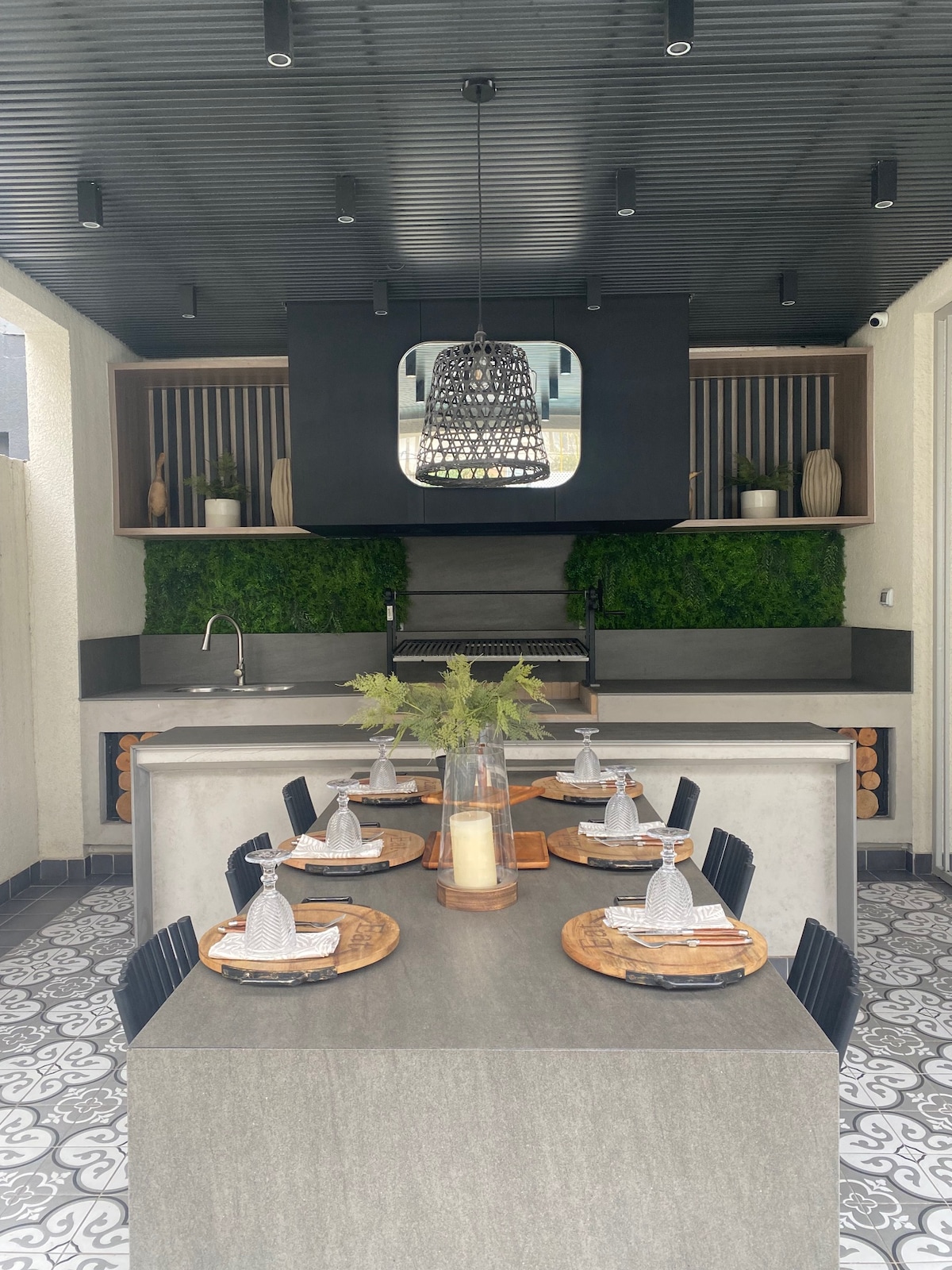 An outdoor dining area is equipped with a long table set with wooden plates, glasses, and elegant napkins. A backdrop of greenery is featured, enhanced by modern lighting fixtures overhead. A grill is positioned in the corner, complemented by a sleek countertop.
