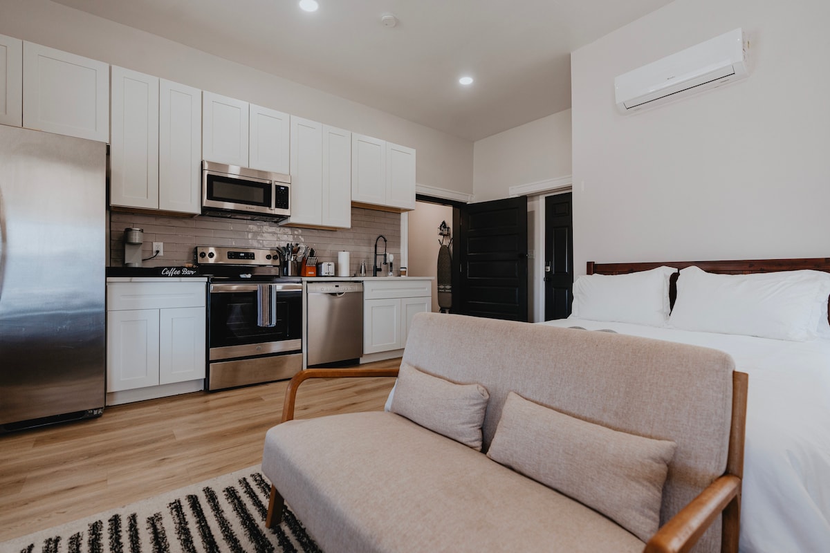 A bright studio space is displayed with a fully equipped kitchen featuring stainless steel appliances and sleek cabinetry. A cozy seating area with a light-colored sofa is positioned in front of the bed, creating an inviting atmosphere. Natural light floods the room.
