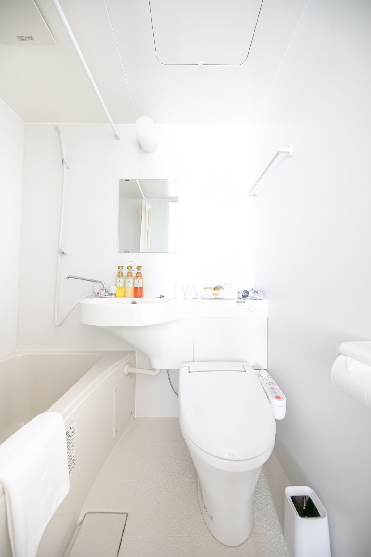 A clean and minimalist bathroom is displayed, featuring a modern toilet with a control panel. A sink with toiletries is positioned next to a small mirror. The bathtub is partially visible, and light colors enhance the brightness of the space.