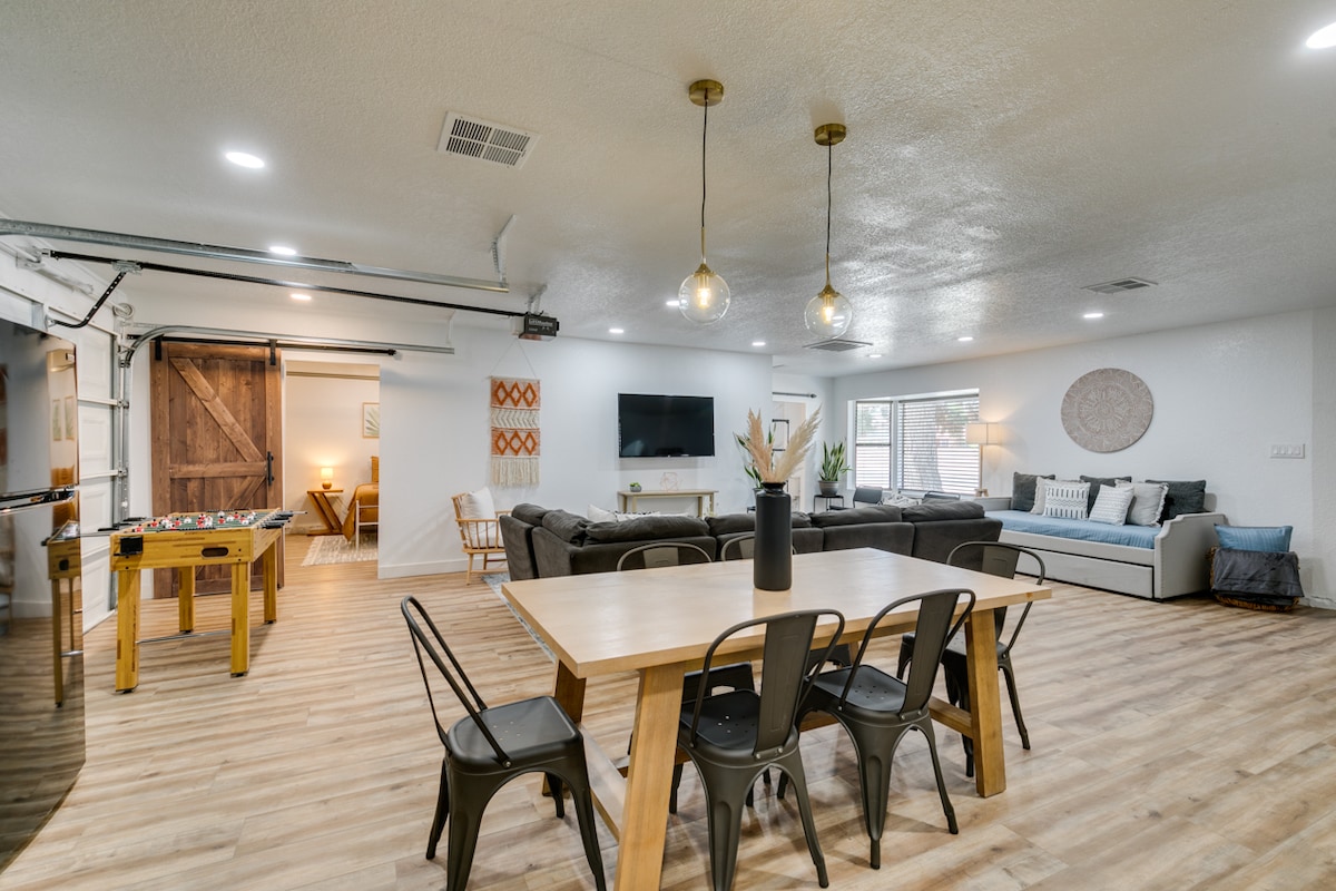 An open concept living area features a spacious dining table surrounded by black chairs. A comfortable seating arrangement with a large sectional sofa and a mounted TV is visible. Natural light streams in through large windows, creating a bright and airy atmosphere.