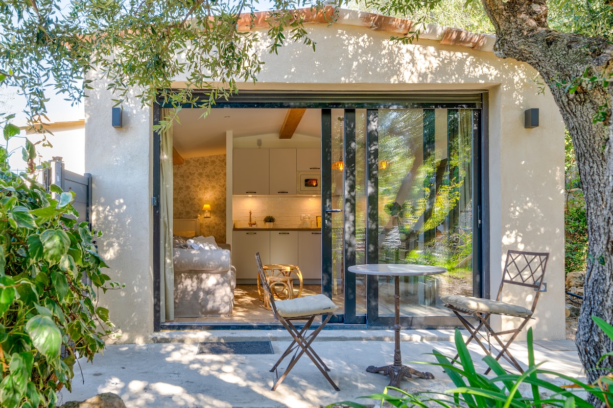 An outdoor seating area features a small round table and two folding chairs, positioned near the entrance. Large glass doors lead to a well-lit interior, revealing a cozy living space with modern kitchen amenities, surrounded by lush greenery.