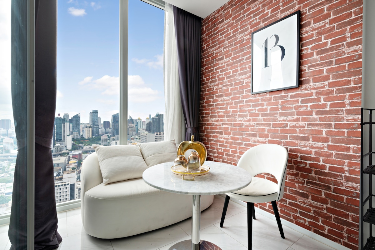 A cozy corner features a round marble table accompanied by a stylish white chair. A curved white sofa provides additional seating, with a striking brick wall serving as a backdrop. Large windows showcase a panoramic view of the city skyline, allowing natural light to fill the space.