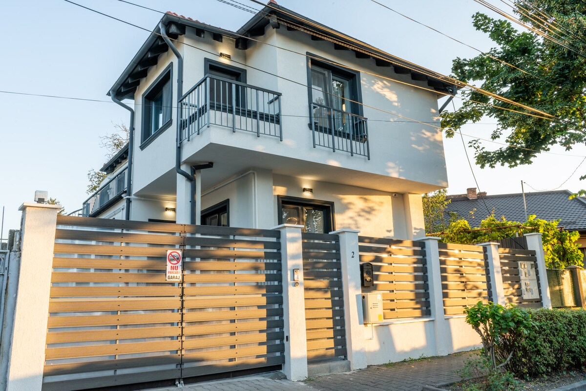 An exterior view of a two-story house is presented, featuring a gated entrance with horizontal slats. The façade displays large windows, black railings on balconies, and subtle outdoor lighting. Surrounding greenery adds a touch of nature to the urban setting.