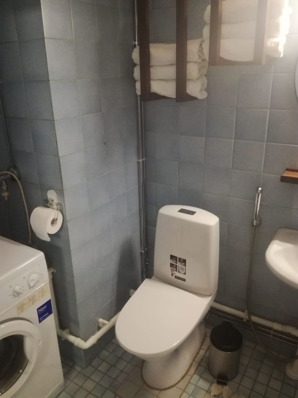 A functional bathroom space features light blue tiled walls, a washing machine, and a white toilet. Fresh towels are neatly arranged on a nearby shelf. A round mirror hangs above a small sink, illuminated by overhead lighting.