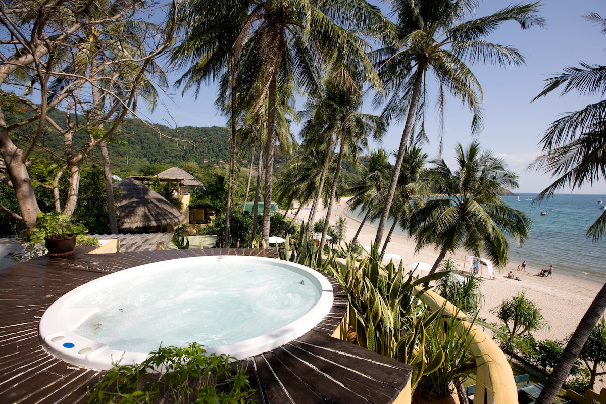 A private hot tub is positioned on a wooden deck surrounded by tropical plants, offering views of a sandy beach and clear blue water. Palm trees sway gently in the breeze, enhancing the tranquil outdoor setting by the water's edge.