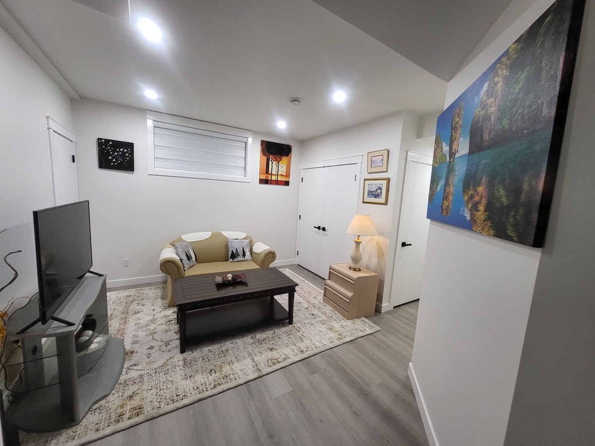 A cozy living area features a comfortable sofa and a coffee table, with a television positioned on a stand. Soft lighting is provided by ceiling-mounted lights and a lamp beside a dresser. Art adorns the walls, contributing to a relaxed atmosphere.
