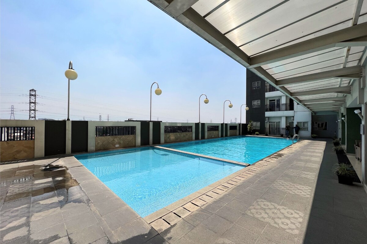 An outdoor swimming pool is featured underneath a spacious covered area, with lounge lighting installed along the perimeter. Clear blue water reflects the surrounding sky, and the pool deck is lined with potted plants, providing a refreshing space for relaxation and leisure.