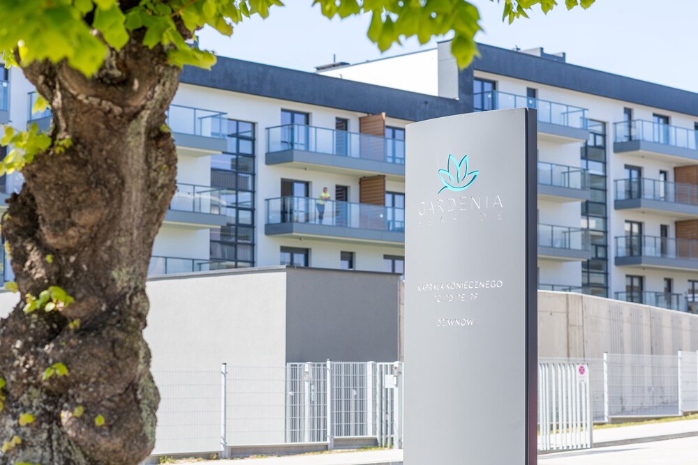 A modern building complex is presented with a sleek sign displaying the name 'Gardenia Seaside.' Balconies are visible on the upper floors, surrounded by greenery. A pathway leads to a gated entrance, enhancing accessibility and secure access to the property.