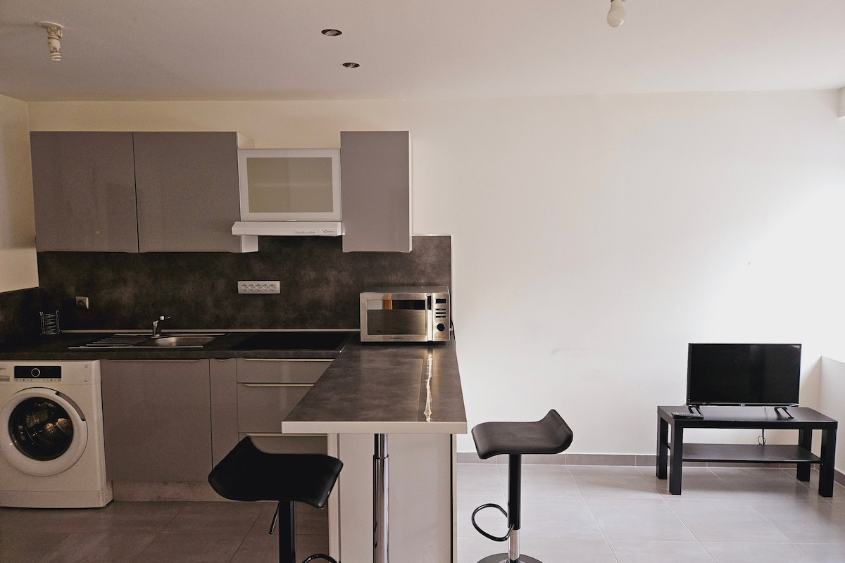 A modern kitchen space features sleek grey cabinetry and a functional countertop. A washing machine is situated beside the sink. Bar stools accompany the kitchen island, and a compact dining area contains a small TV on a stand against a wall.