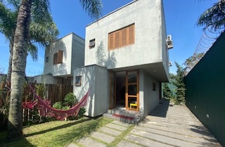 A modern two-story house is set within a surrounding garden. A pathway leads to the entrance, while a hammock is visible among the greenery. Large windows and wooden shutters enhance the exterior, and ample sunlight illuminates the building's minimalist design.