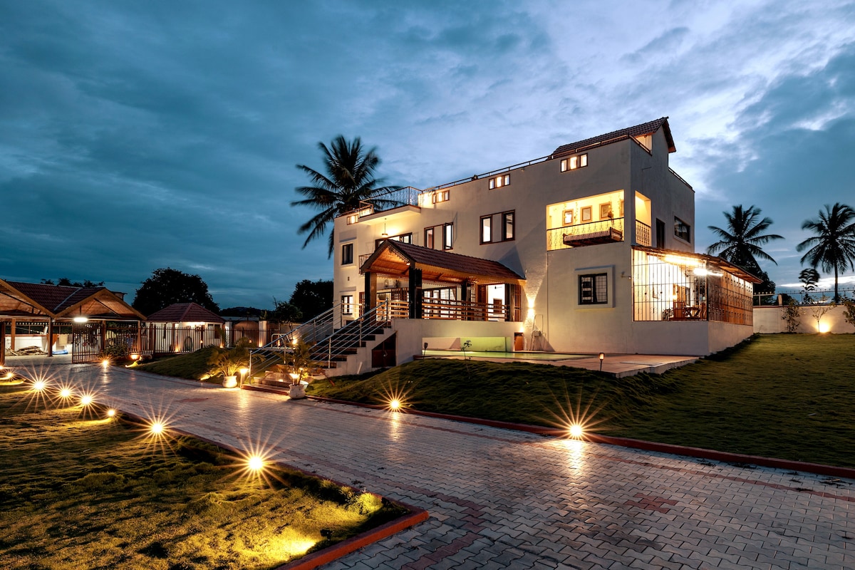 The exterior of the resort is viewed during twilight, showcasing a spacious building with a warm wooden entrance and multiple windows. Pathway lights illuminate the surrounding landscape, highlighting the lush green lawn and palm trees in the background.