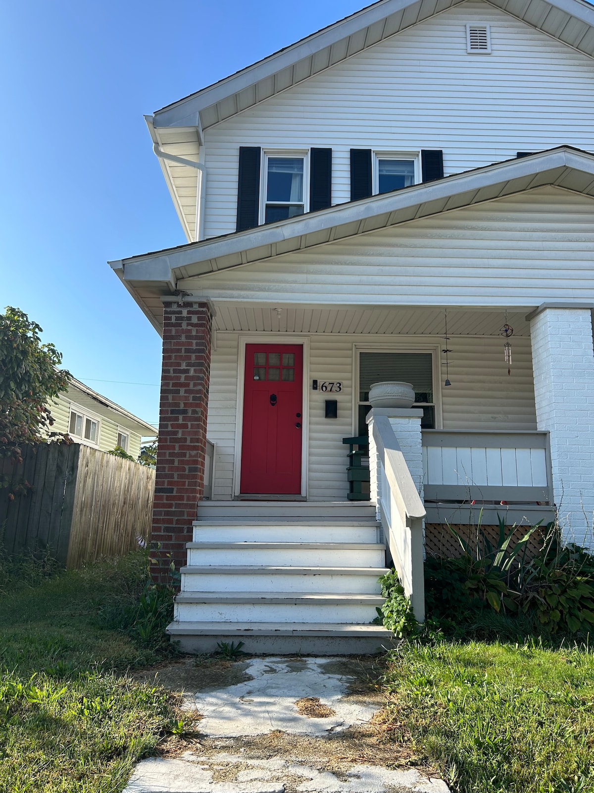 The exterior of the house showcases a bright red front door, contrasting with white siding and black shutters. A small porch is visible, with a single chair positioned beside the entrance. Steps lead up to the door, framed by a lush green lawn and surrounding garden.