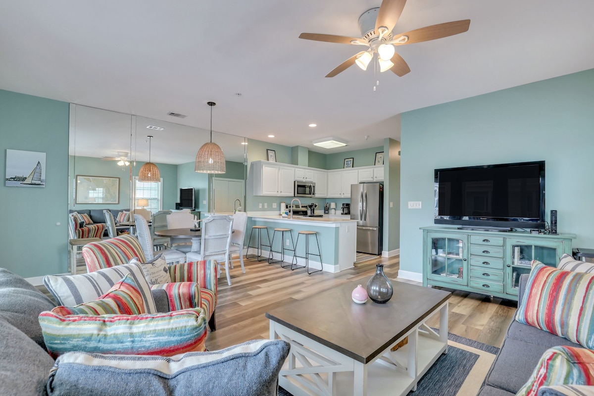 An open-concept living area showcases a modern kitchen with stainless steel appliances and a spacious island. Cozy seating arrangements include a striped couch and a decorative coffee table. Light wood flooring enhances the airy atmosphere, complemented by soft blue walls and ceiling fan overhead.