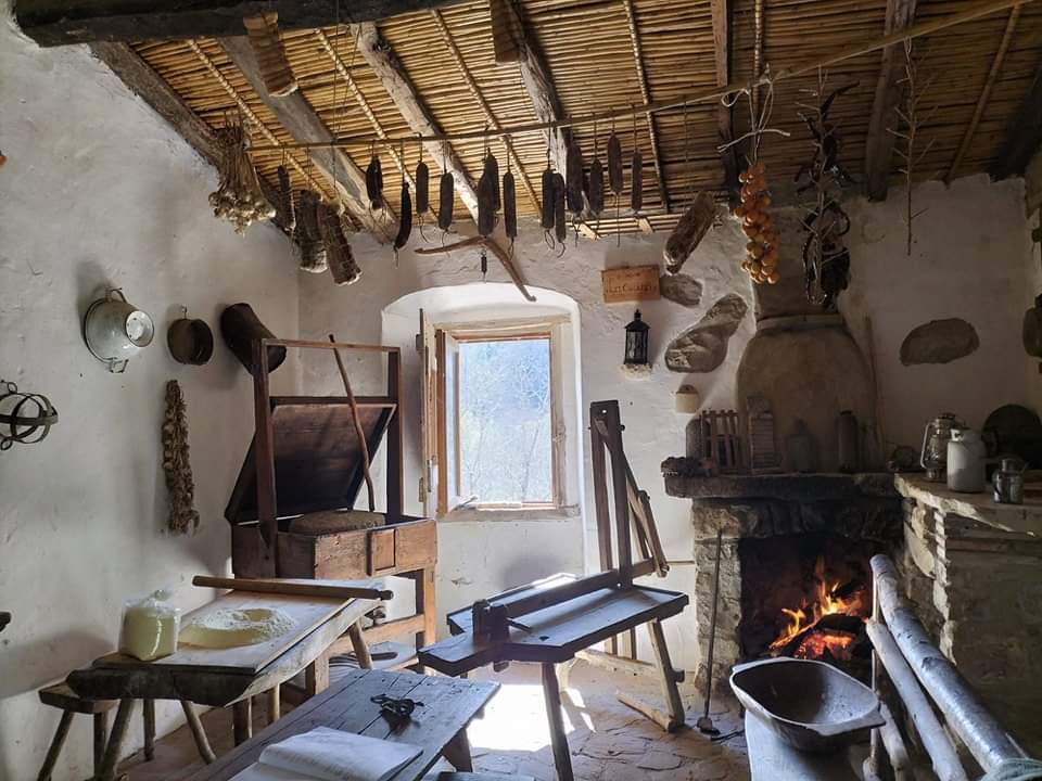 A rustic kitchen space features traditional wooden furniture, including tables and a workbench. Dried herbs and vegetables hang from the ceiling, while a cozy fire burns in a stone fireplace. Natural light enters through a window, illuminating the earthy tones of the walls.