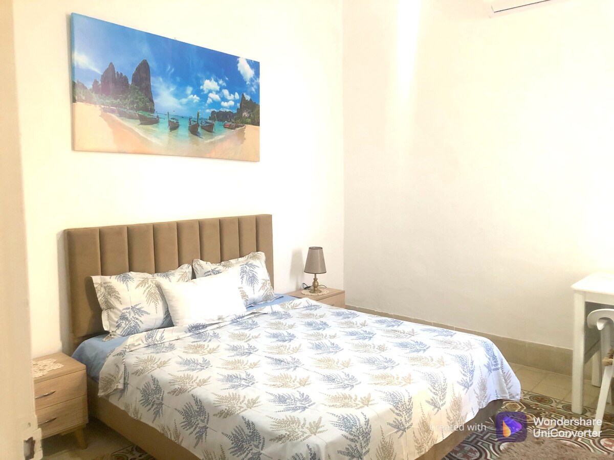 A bedroom features a spacious bed adorned with a light blue floral duvet cover. A soft lamp is placed on a bedside table, and a small white desk is visible in the corner. A large artwork above the bed depicts a scenic beach landscape.