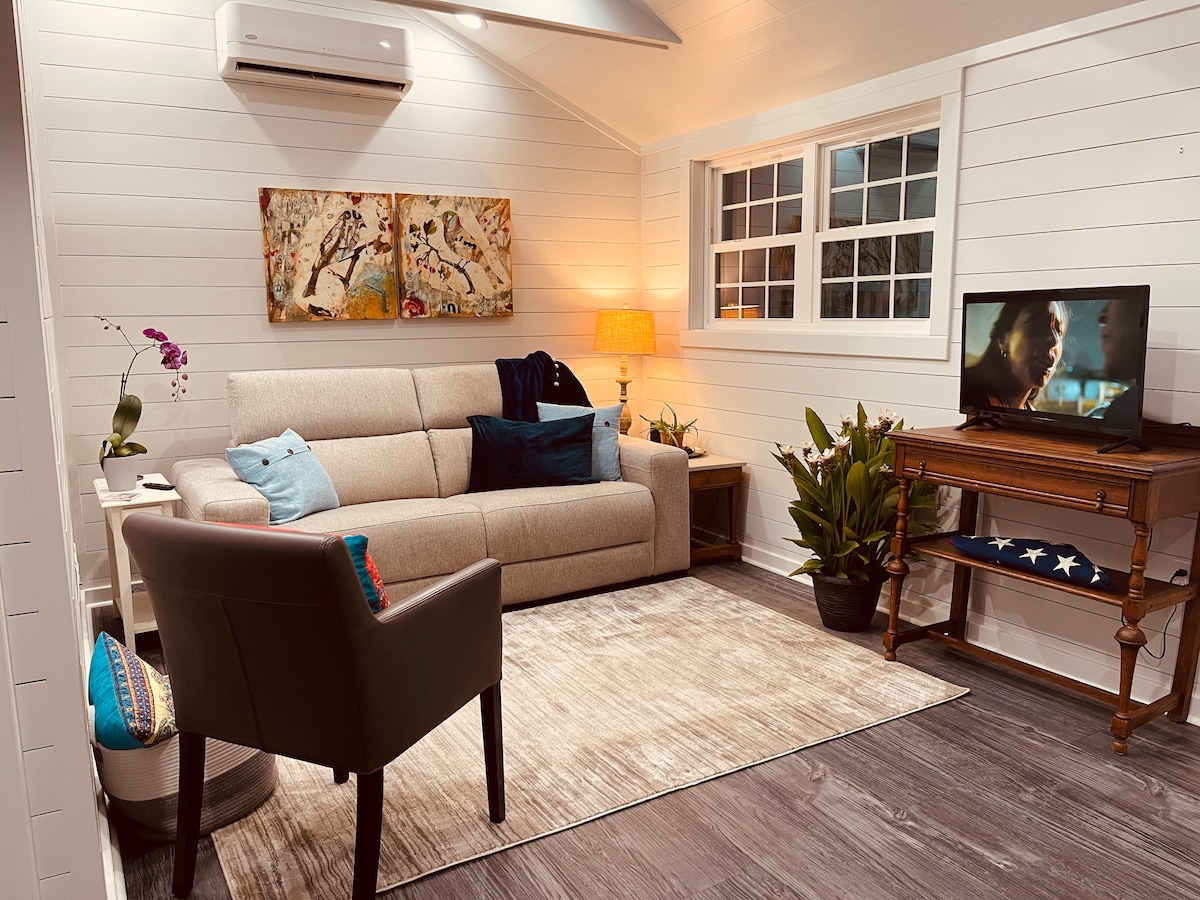 A cozy living area is highlighted by a light-colored sofa adorned with colorful cushions. A wooden table is positioned beside the sofa, while a small television sits atop a vintage-style stand. Indoor plants and artwork bring a touch of natural warmth to the space.