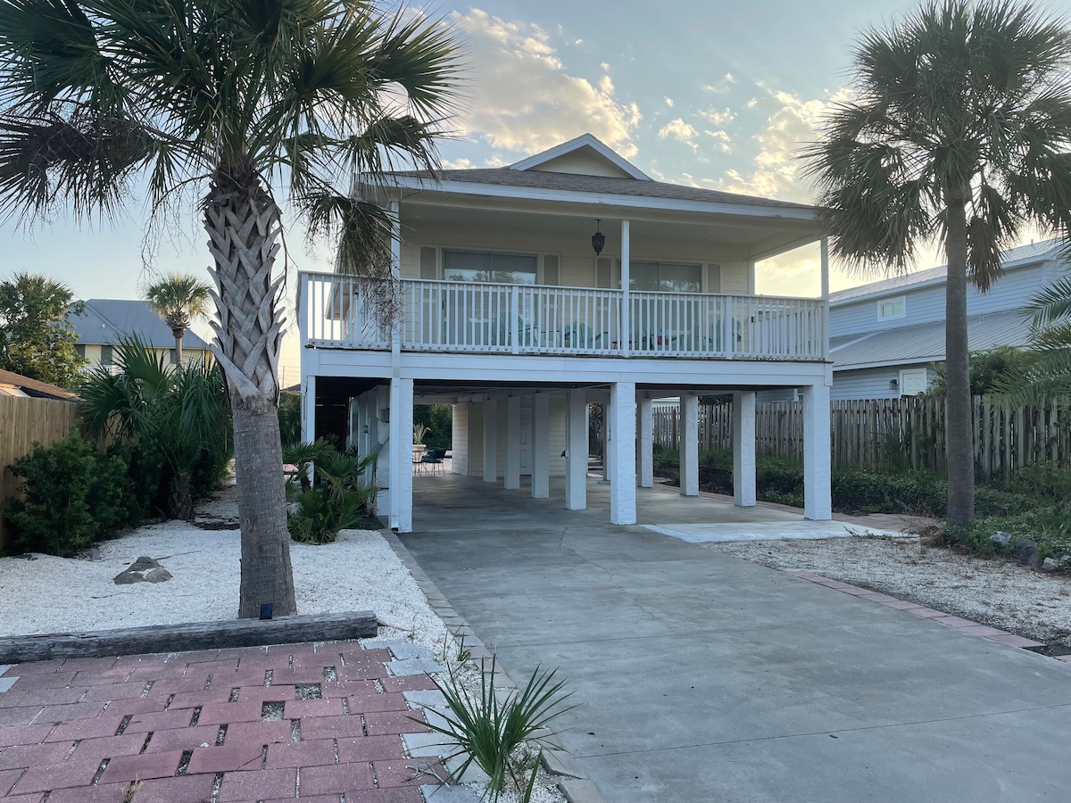 A two-story beach house is showcased amid palm trees and neatly landscaped greenery. The home features a spacious deck with white railings, supported by white columns, and is set on a concrete driveway with decorative landscaping in the foreground.