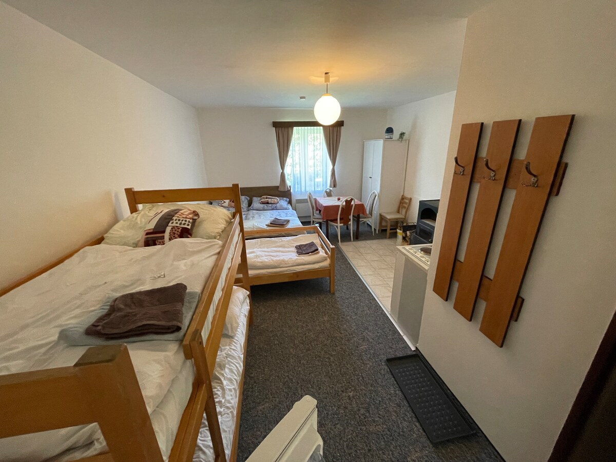 A spacious room features wooden bunk beds and a separate sleeping area with additional beds. A table and chairs are positioned near a window, while a kitchenette is visible in the corner. Coat hooks are mounted on the wall for convenience.