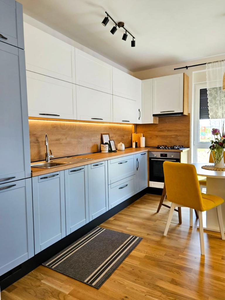 A modern kitchen features blue and white cabinetry with wooden accents. A stainless steel sink and stove are positioned next to a small dining table, accompanied by a yellow chair. The space is illuminated by overhead lighting, and a soft rug adds warmth to the wooden floor.
