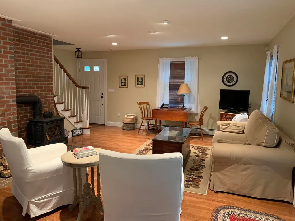 A spacious living area features hardwood floors and a cozy seating arrangement with light-colored chairs and a sofa. A wooden table serves as a work space, while a flat-screen television is positioned nearby. Natural light filters in through the windows, enhancing the warm ambiance.