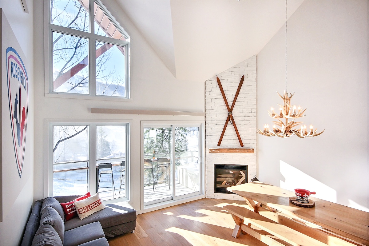 A bright living area features large windows allowing natural light to fill the space. A modern fireplace is complemented by wooden accents and a stylish chandelier. A spacious dining table sits adjacent to a comfortable sofa, creating a welcoming environment for gatherings.